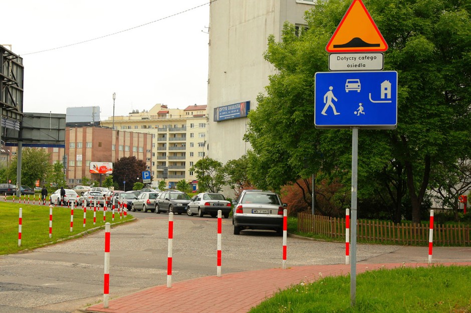 Strefa zamieszkania (znak D-40): piesi mają pierwszeństwo, a parkować wolno tylko w wyznaczonych miejscach, czyli zupełnie inaczej niż na tym zdjęciu. Informacja o progach zwalniających jest tu zbędna, bo w strefie zamieszkania można je ustawiać bez ostrzeżenia. 