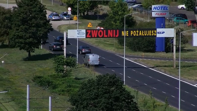Znak nawołujący do zwolnienia a za nim patrol policji