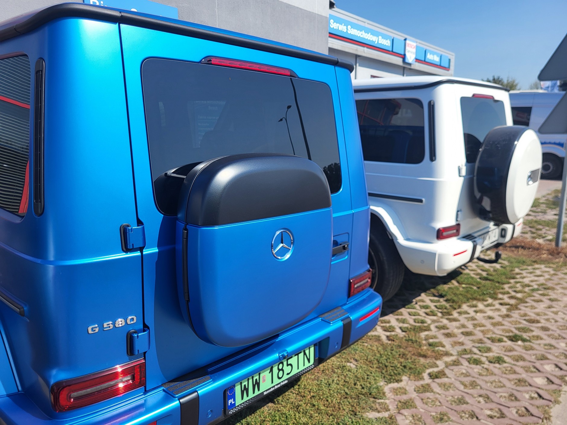Nie, elektryczny Mercedes G580 EQ nie ma na tylnej klapie kwadratowej dojazdówki – to schowek na kable! Na drugim planie wersja spalinowa z kołem zapasowym.